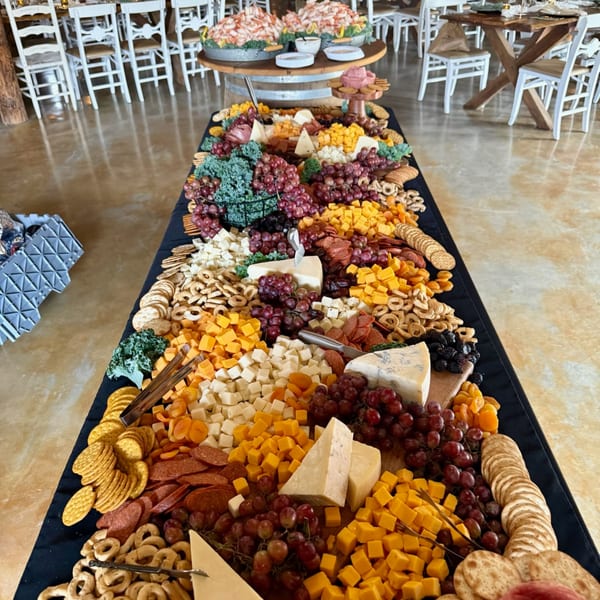 A long deli charcuterie board overflowing with cheeses, meats, grapes, crackers, and dried fruit.