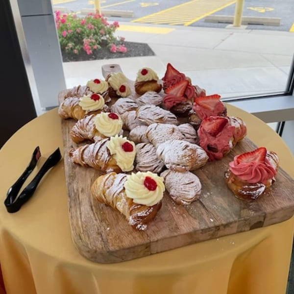 A wooden board filled with Rocco's Market & Deli pastries, some with cream and cherries, others with strawberries.