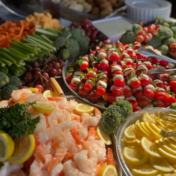 A deli catering spread with shrimp, caprese skewers, vegetables, and lemon slices.