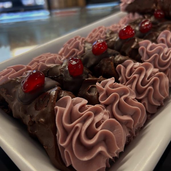 Chocolate-dipped cannoli with pink frosting and candied cherries on a white platter.