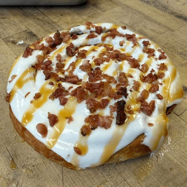 A large cinnamon roll topped with white icing, caramel drizzle, and crispy bacon bits on a wooden surface.