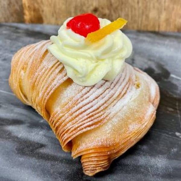 A Rocco's Market & Deli sfogliatella pastry, dusted with powdered sugar and topped with cream, a cherry, and orange peel.