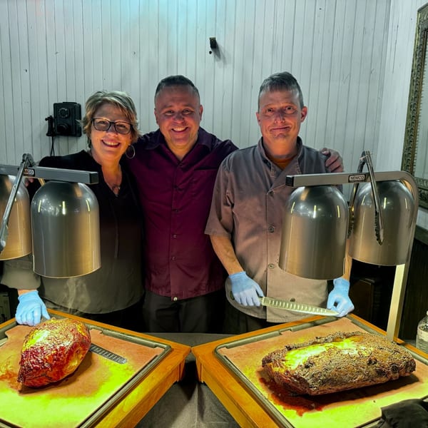 Three Rocco's Market & Deli staff members smiling behind two large roasts under heat lamps.