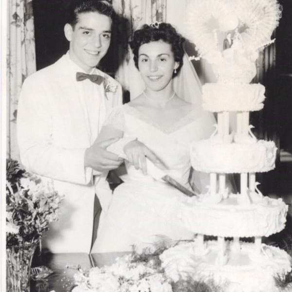 A black and white photo of a smiling bride and groom cutting a multi-tiered wedding cake.