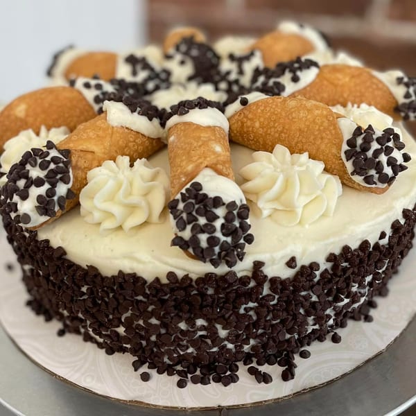 A Rocco's Market & Deli cannoli cake with white frosting, mini cannolis, and chocolate chips.