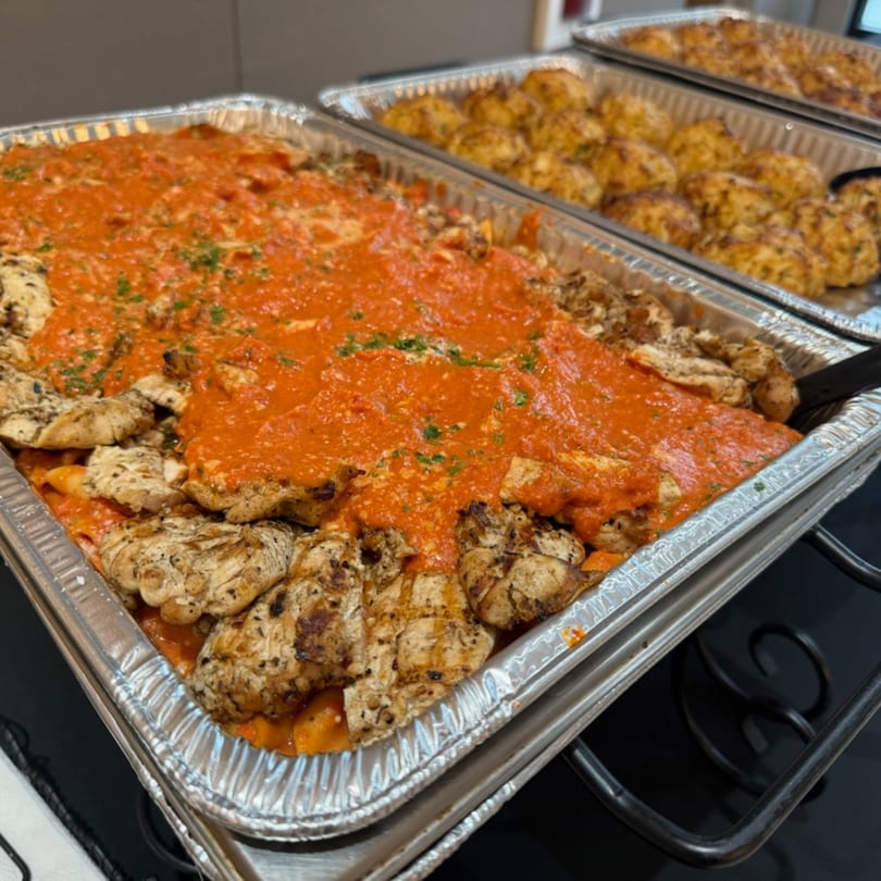 Two foil pans of deli food, one with grilled chicken and red sauce, the other with golden-brown items.